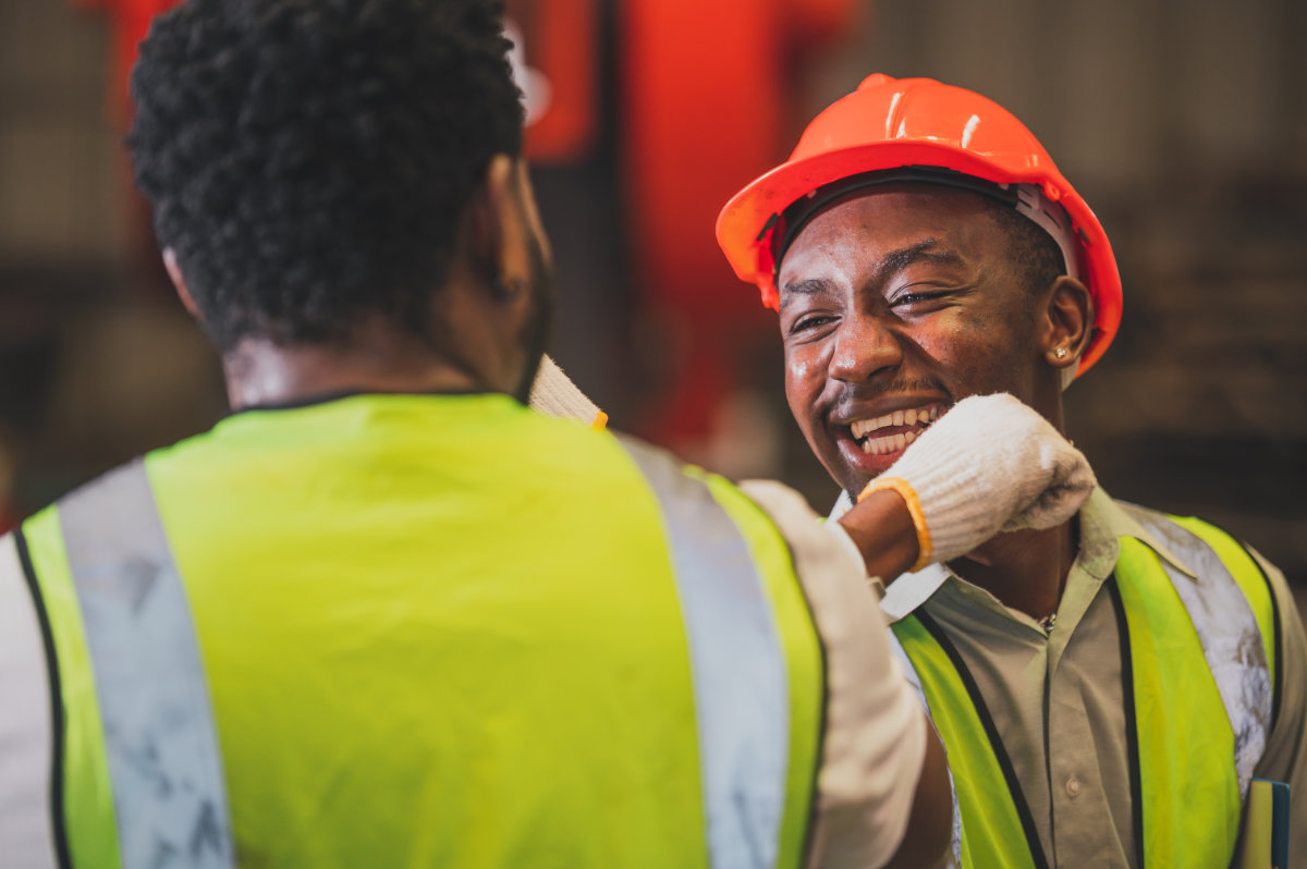 Worker in orange hard hat laughing with a colleague in a yellow safety vest at an industrial or construction site.