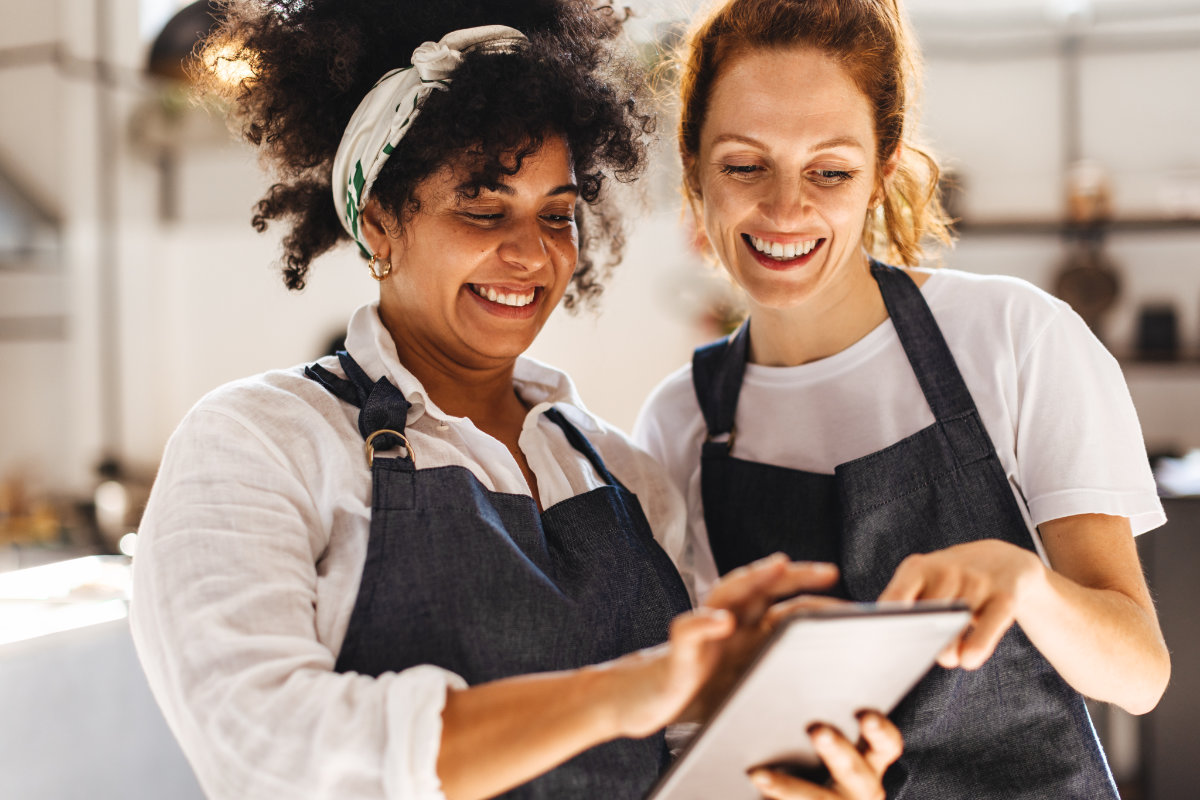 Two smiling women wearing denim aprons looking at a tablet together, appearing to work in a small business or restaurant setting.